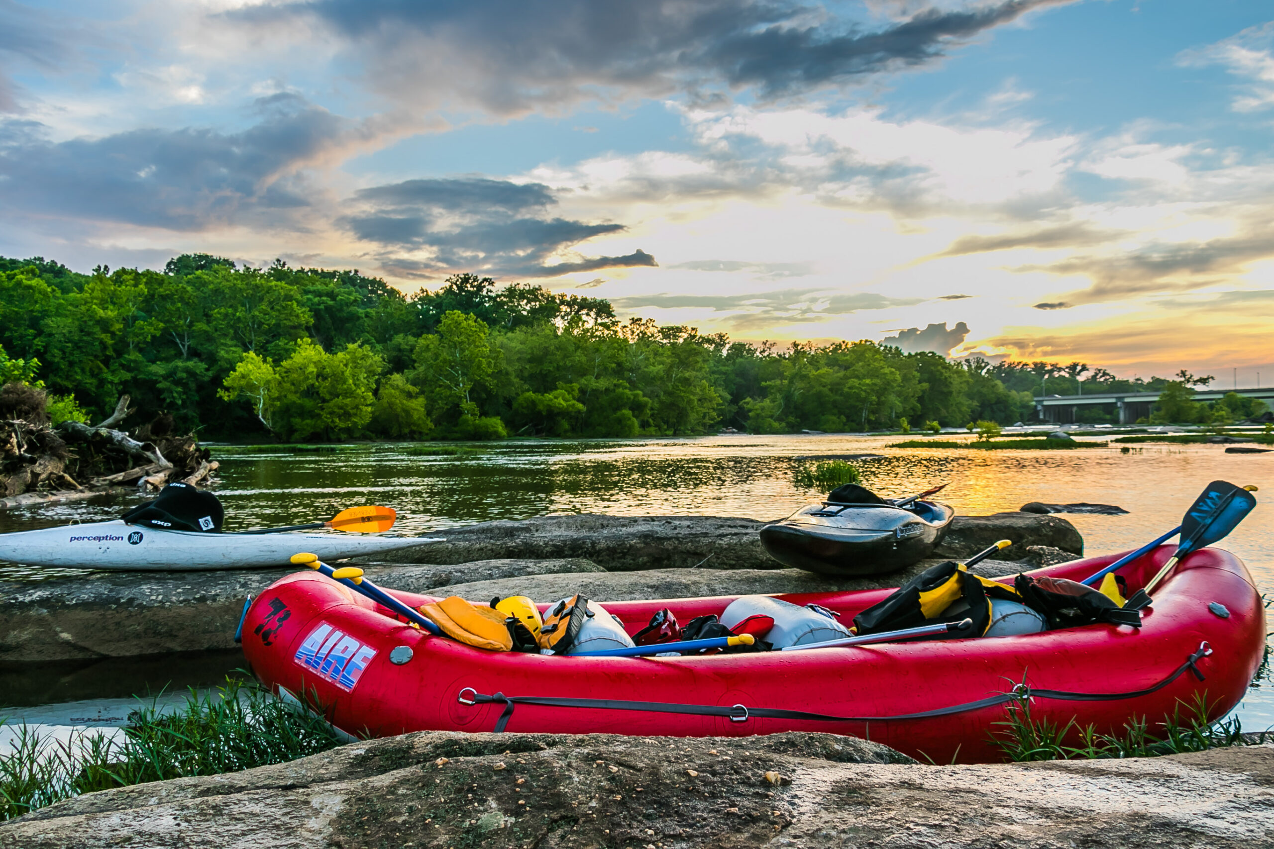 A red inflatable raft loaded with paddles and life jackets, pulled up on flat rocks beside the James River at sunset, with two kayaks resting nearby