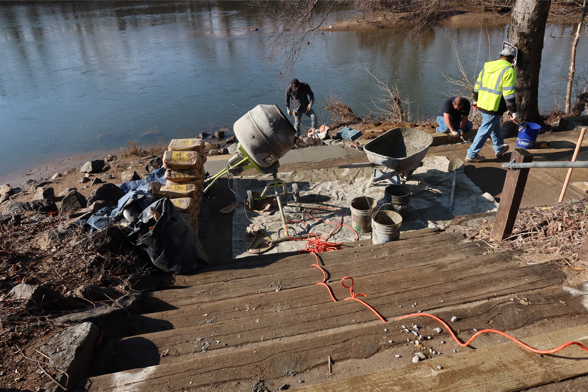 Three volunteers pouring and finishing a new concrete walkway beside the James River at the 14th Street Takeout, with a cement mixer, wheelbarrow, and wooden steps in the foreground